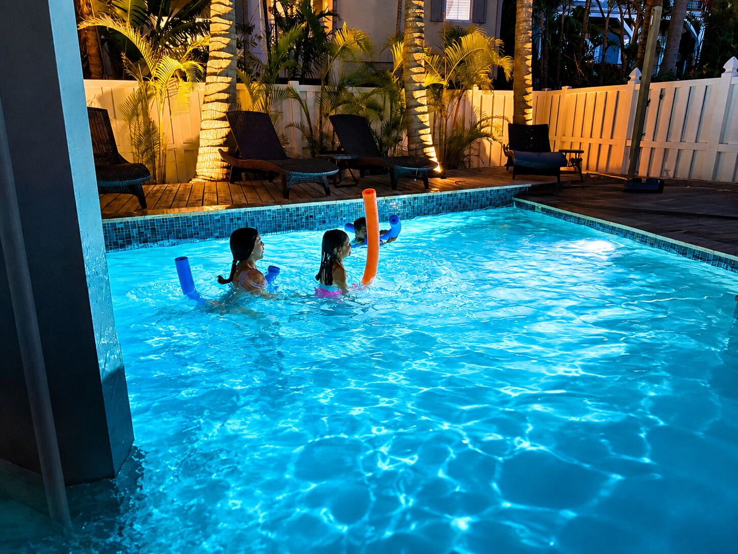 Ein Schwimmbecken mit zwei spielenden Kindern, umgeben von Liegestühlen und Palmen.