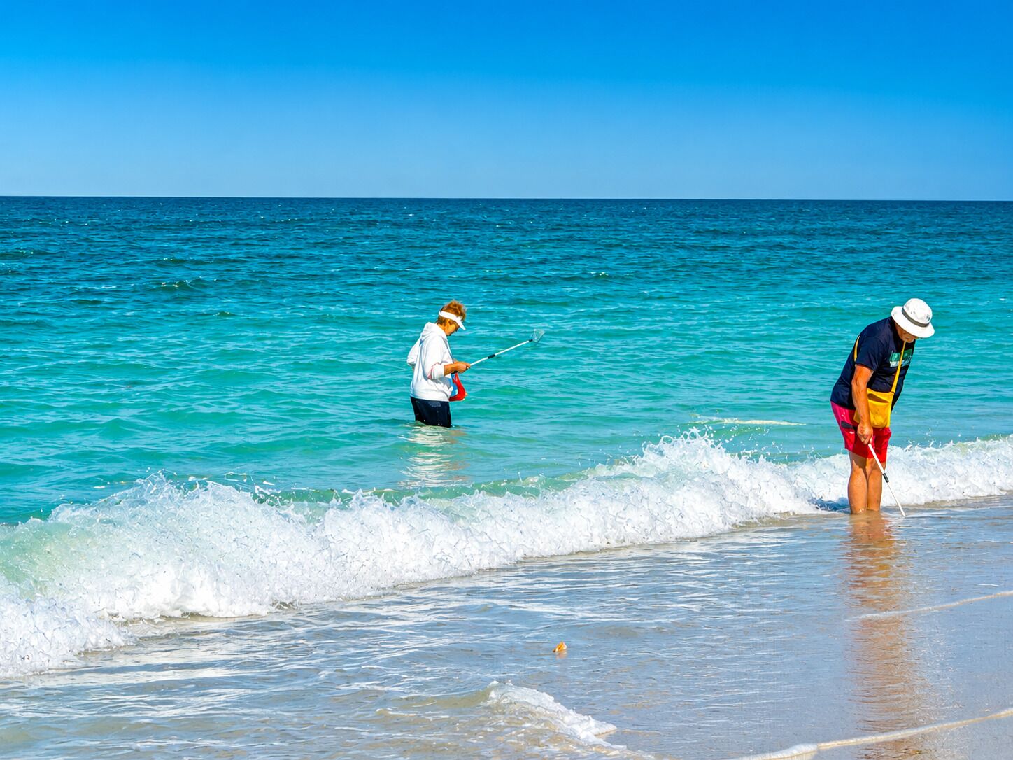 Two individuals fishing on a beach with clear turquoise water and a calm sea.