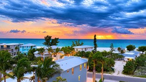A coastal resort with multiple buildings, palm trees, and a view of the ocean at sunset. - Shore to Please | Gulf Views 🌊 Elevator | Beach 1 Min 🌴 | Heated Pool & Spa 🌅 (Anna Maria)