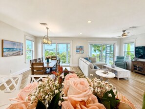 A spacious living room with a dining area, large windows, and a ceiling fan. - Shore to Please | Gulf Views 🌊 Elevator | Beach 1 Min 🌴 | Heated Pool & Spa 🌅 (Anna Maria)