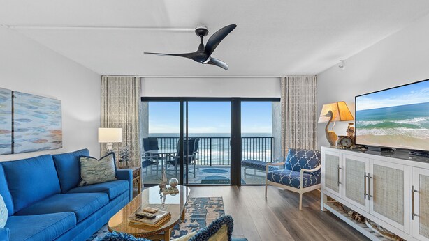 A modern living room with a blue sofa, a coffee table, a television, and a view of the ocean through large glass doors.