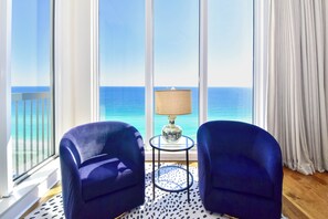 A room with a large window offering a view of the ocean, two blue chairs, a small round table with a lamp, and a patterned rug. - Spacious Beachfront Penthouse w/ Stunning Views & Great Amenities! (Destin)
