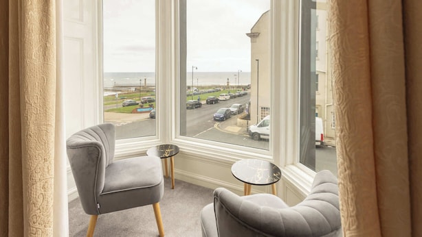 A room with two grey armchairs and a small round table, a large window with a view of a parking lot and buildings. - Host & Stay | Glaisdale at the Sandbeck (Whitby)