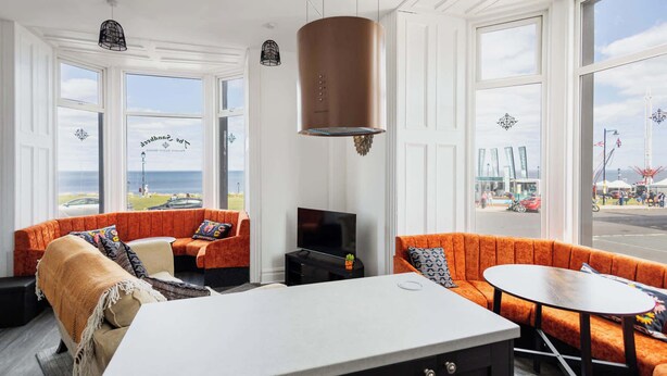 Ein modernes Hotelzimmer mit einem großen Fenster, einer zentralen Sitzecke mit orangefarbenen Sofas, einem Esstisch und einer Küchenzeile.