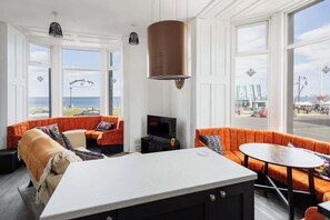 Et moderne hotelværelse med et stort vindue, et centralt opholdsområde med orange sofaer, et spisebord og et tekøkken.