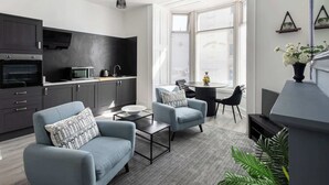 Un salon moderne avec des canapés gris, une table basse et un téléviseur. On y trouve également un coin cuisine avec des meubles sombres et un four à micro-ondes. La pièce est dotée de grandes fenêtres équipées de stores, et une plante est visible sur le côté droit.
