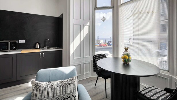 Une cuisine moderne avec un plan de travail noir, un four à micro-ondes et un évier. On y trouve un coin repas avec une table ronde et des chaises, et une vue sur la ville grâce à de grandes fenêtres.