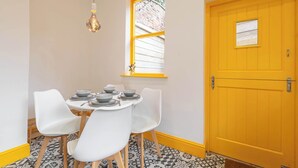 Un comedor con una mesa dispuesta para cuatro, sillas blancas, una lámpara colgante, un ventanal con marco amarillo y una puerta amarilla.