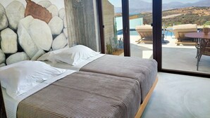 Ett sovrum med en vägg av natursten, en säng med vita sängkläder och ett mönstrat överkast, och utsikt över en pool och ett utomhusområde med sittplatser.