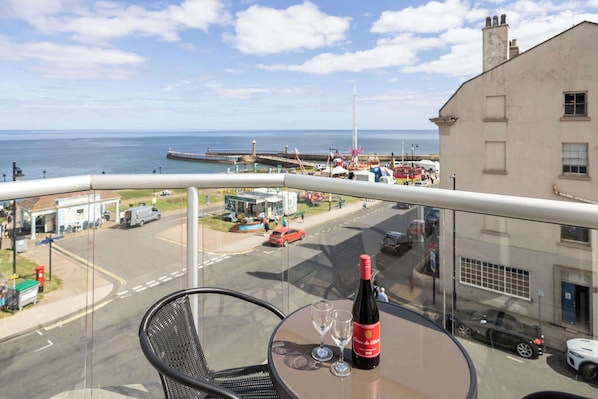 A balcony with a table, a bottle of wine, and two glasses. The view includes a coastal area with a pier, buildings, and parked cars.