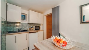 A compact kitchen with white cabinets, a microwave, and a sink. There's a wooden door and a white basket with fruits on the counter.