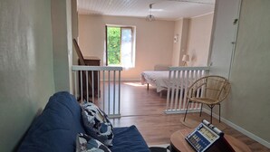 Ein Zimmer mit einem blauen Sofa, einem Korbstuhl, einem kleinen Holztisch mit einem Buch, einer weißen Geländer und einem Bett im Hintergrund.
