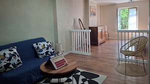 Ein Wohnzimmer mit einem blauen Sofa, einem kleinen runden Tisch, einer weißen Geländer und einem Korbstuhl, mit Blick auf einen Balkon mit einem Fenster.