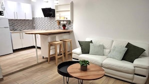 Eine moderne Küche mit weißen Schränken, einer gefliesten Spritzschutzwand und Pendelleuchten. Es gibt ein weißes Sofa mit grünen Kissen und einen kleinen runden Tisch mit einer Pflanze.