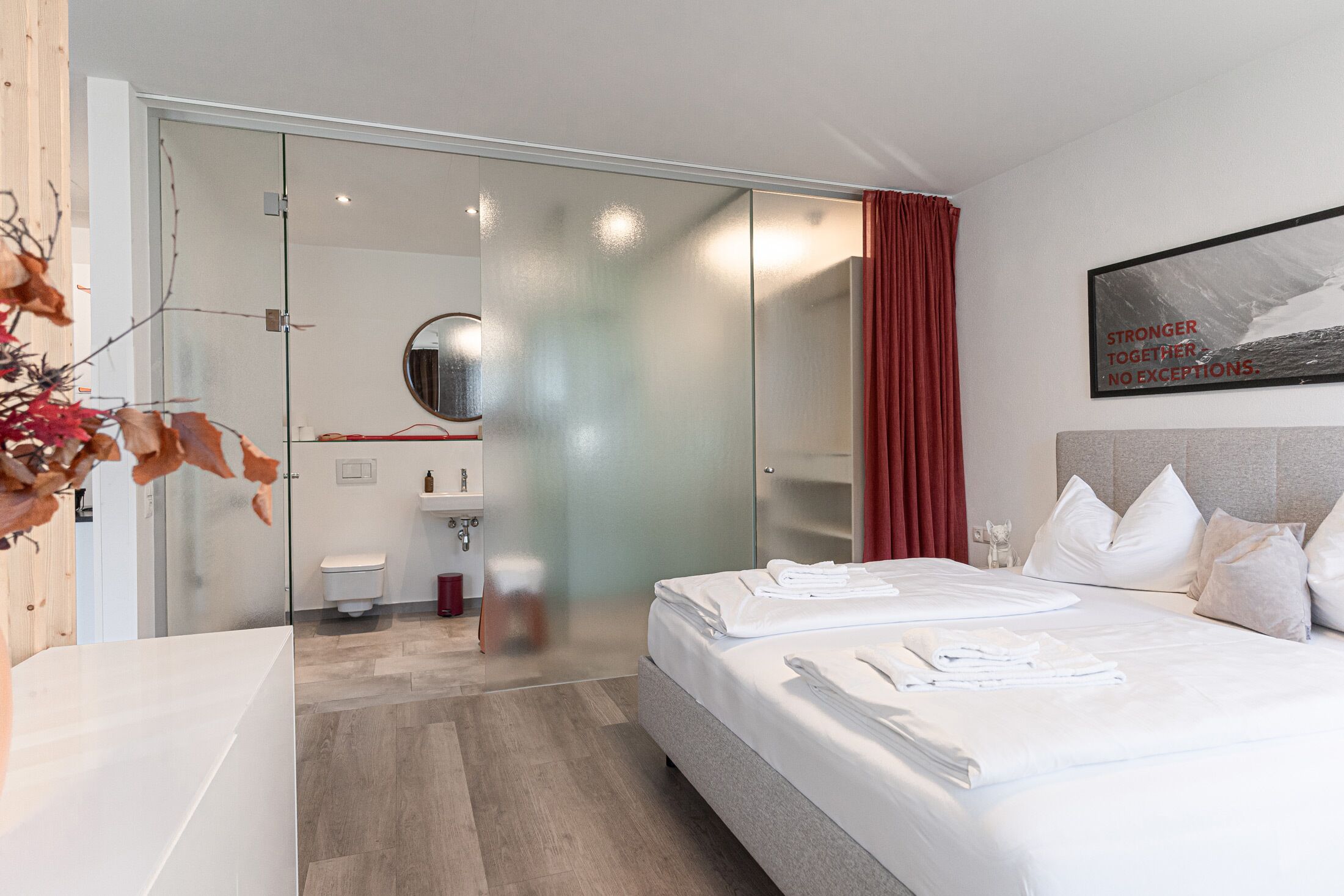 A modern hotel room with a large bed, a glass-enclosed shower area, a bathroom with a toilet and sink, and a motivational quote on the wall.
