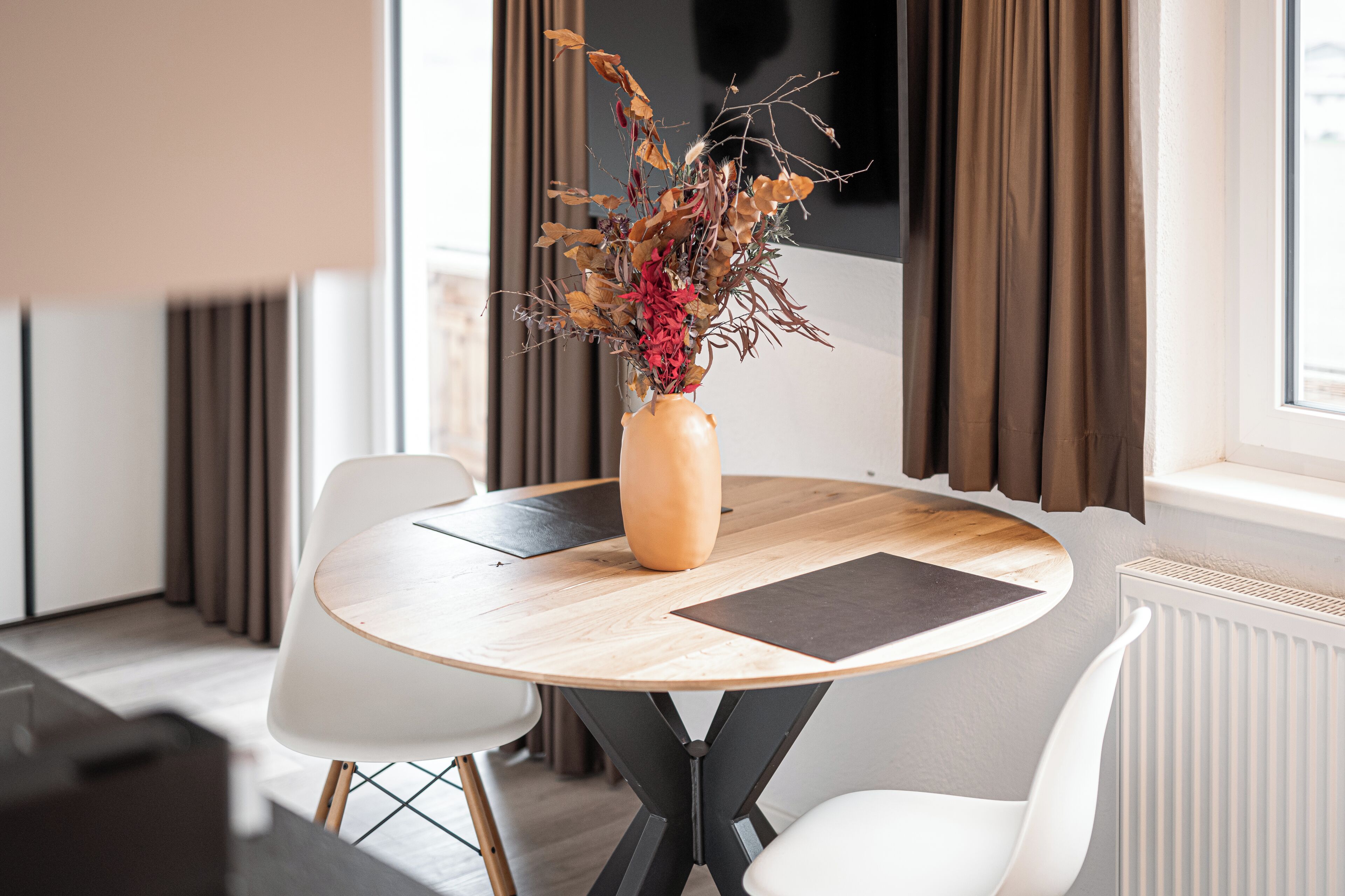 Une table ronde en bois, ornée d’un vase de fleurs séchées, entourée de chaises blanches, et donnant sur une fenêtre avec des rideaux.