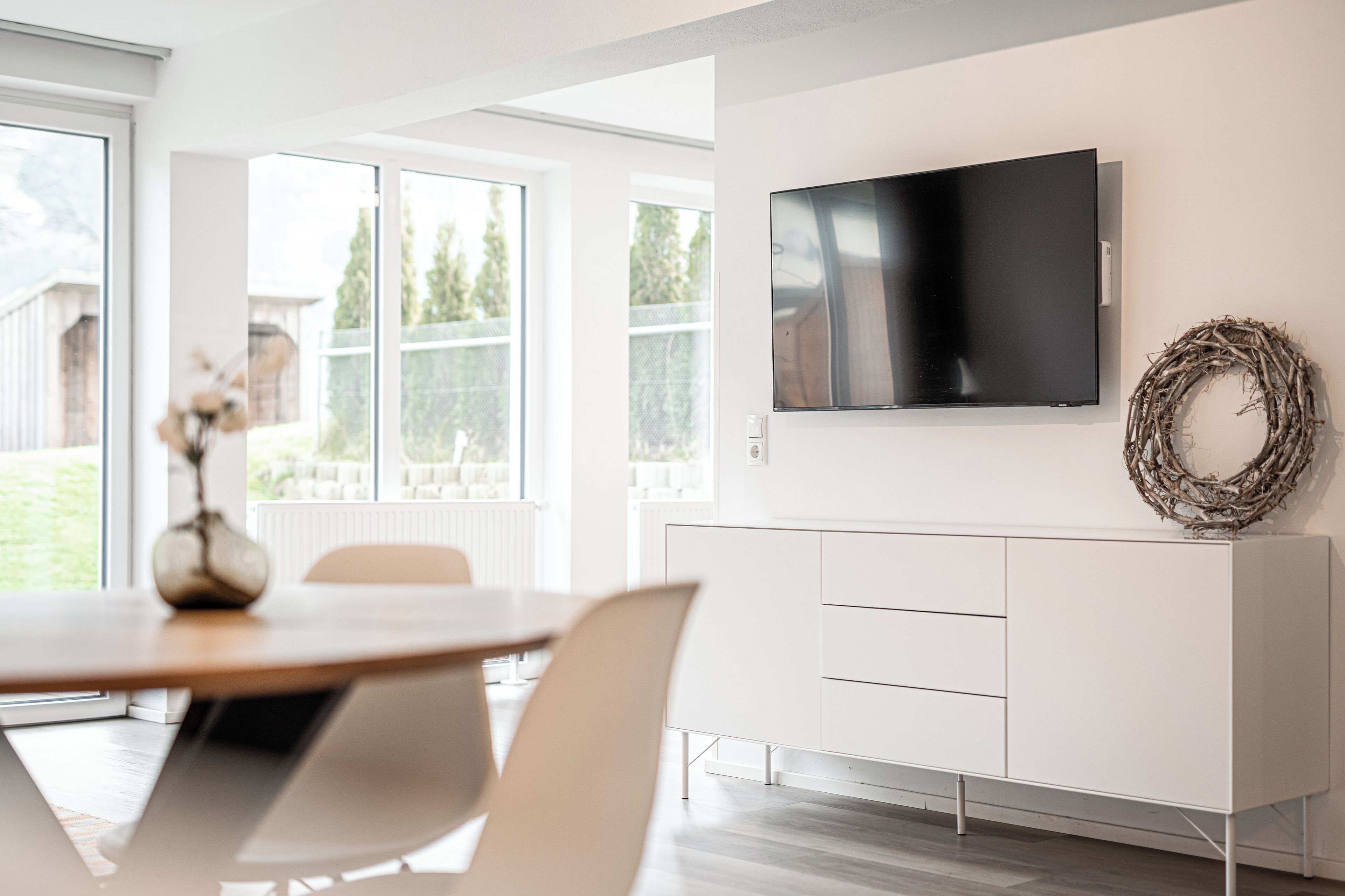 Una sala de estar moderna con un televisor de pantalla plana montado en la pared, un mueble blanco y un comedor con mesa y sillas.