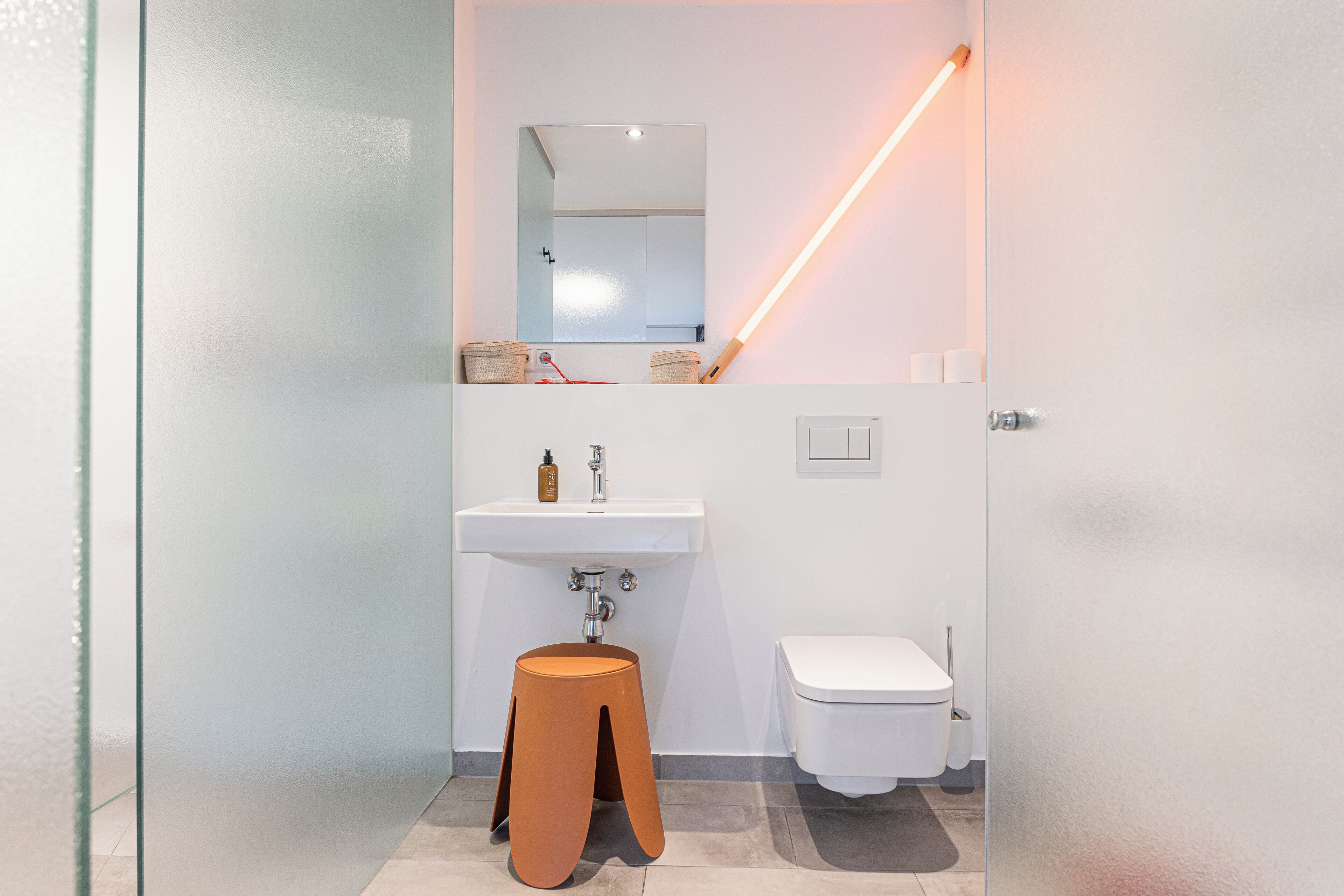 Ein modernes Badezimmer mit Glasabtrennung, einem weißen Waschbecken, einer Toilette und einem Hocker.