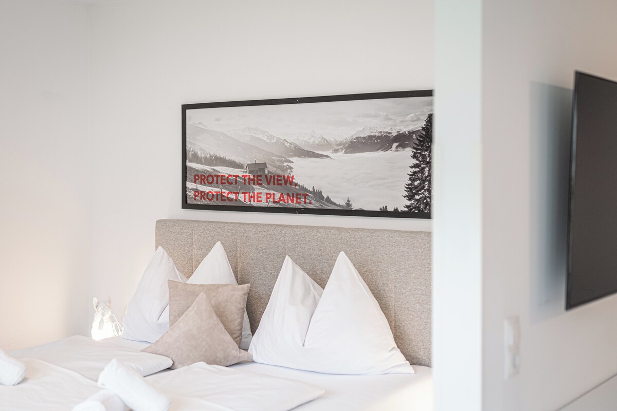 A neatly made bed with white and beige pillows, a framed picture on the wall, and a flat-screen TV mounted on the wall.