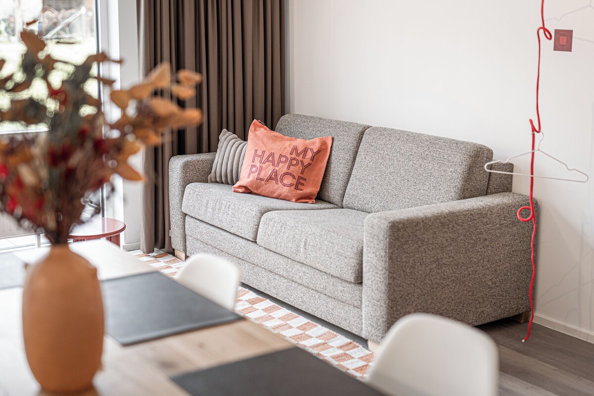 A modern living room with a grey sofa, a decorative pillow, a vase with dried flowers, and a red hanger on the wall.