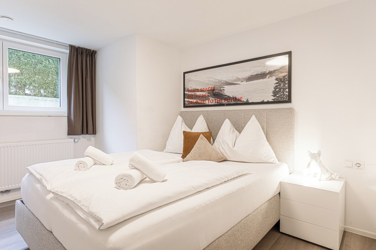 A neatly made bed with white linens and pillows, a bedside table with a lamp, and a framed picture on the wall.