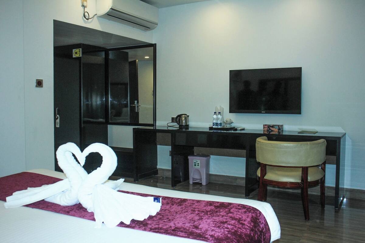 A hotel room with a bed, a desk, a chair, a TV, and a towel artfully arranged on the bed.