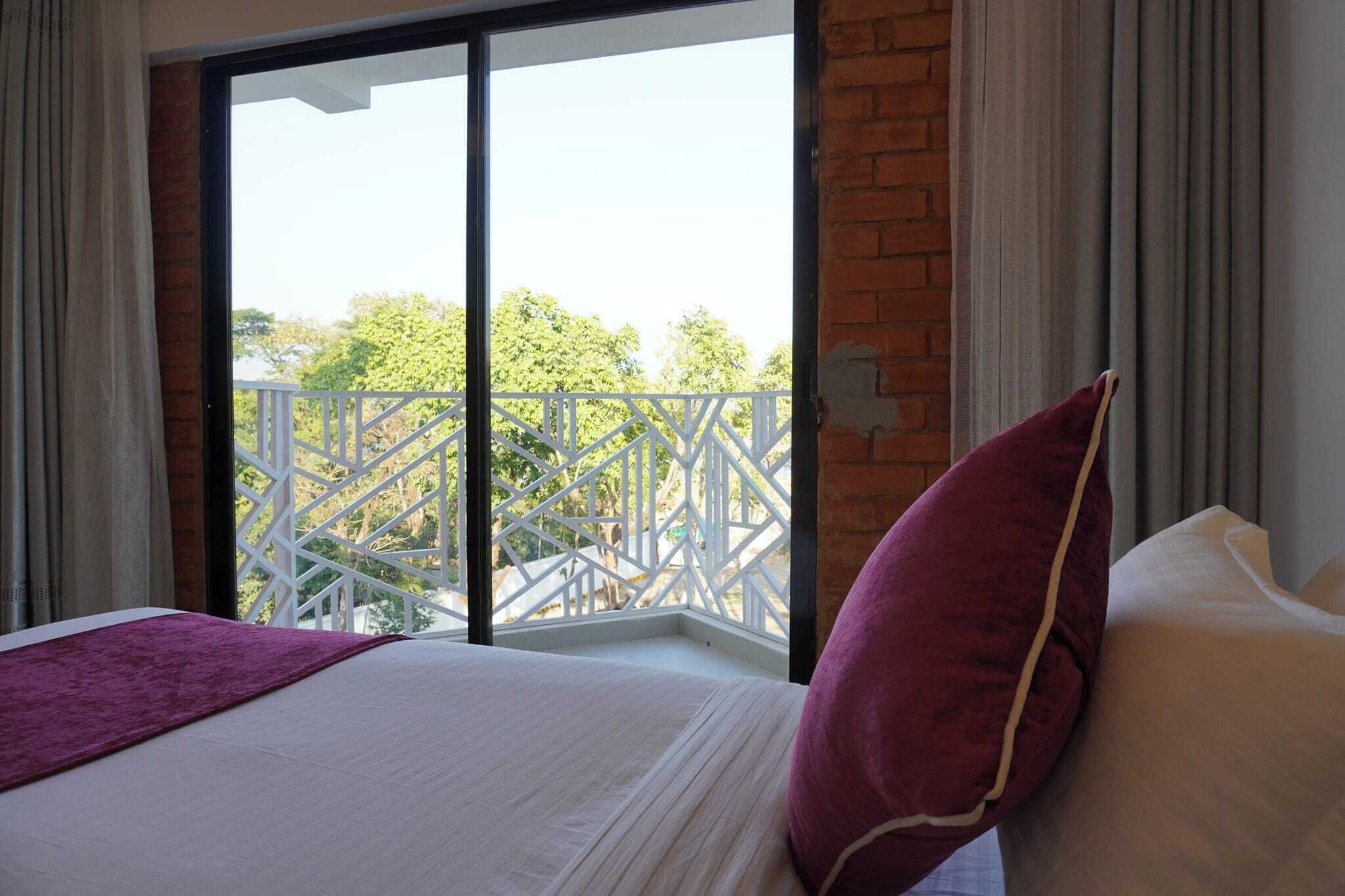 Habitación de hotel con una cama, un cojín morado y vistas a un balcón con barandilla blanca.