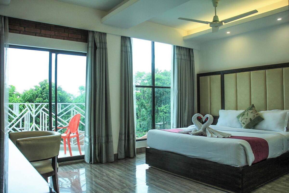 A hotel room with a large bed, a desk, a chair, and a balcony with a view of trees.