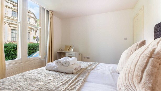 A hotel room with a large bed, a window with curtains, a bedside table with a lamp, and a view of a building outside. - Host & Stay | Flat 3 Club Chambers (Great Malvern)