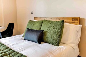 Cama com colchão branco e almofadas verdes texturizadas, cabeceira de madeira e uma pequena mesa com uma cadeira à esquerda.