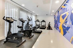 Ein Fitnessraum mit unterschiedlichen Trainingsgeräten, einem großen Fenster inklusive Jalousien sowie einem Wandgemälde.