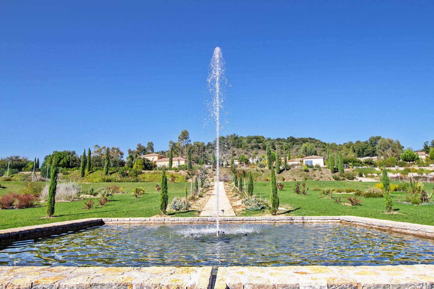 A fountain in a garden with a clear sky, surrounded by greenery and houses in the background.
