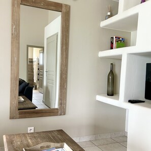 Un salon avec un grand miroir, une table basse en bois et une étagère blanche sur laquelle se trouvent une bouteille et une télécommande.