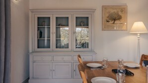 Una mesa de comedor dispuesta con platos, vasos y cubiertos, un aparador blanco con puertas de vidrio y un cuadro en la pared.