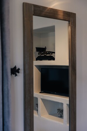 A room with a wooden-framed mirror reflecting a television and shelves with a decorative item and a glass.