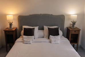 Cama bem arrumada com lençóis brancos e duas almofadas decorativas, com duas mesas de cabeceira com abajures e pequenos objetos decorativos.