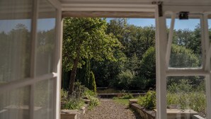 Vista attraverso una finestra su un giardino con vialetto sterrato, panchine in legno e vegetazione rigogliosa.