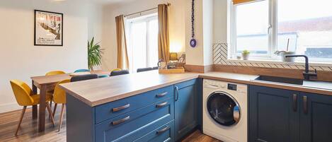 A modern kitchen with a built-in washing machine, wooden flooring, and a dining area with yellow chairs.