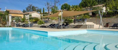 A clear swimming pool with sun loungers and a shaded area, surrounded by a stone wall and greenery.