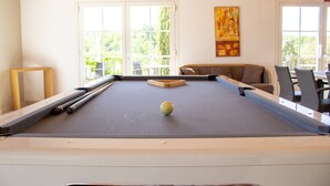 Une table de billard avec une boule blanche, un canapé et une chaise, dans une pièce dotée de grandes fenêtres.