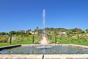 Ein Springbrunnen in einem Garten mit blauem Himmel, umgeben von viel Grün und Häusern im Hintergrund.