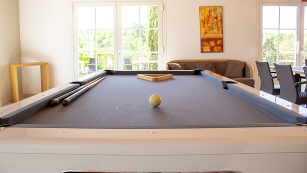 Een biljart met een witte bowlingbal, een bank en een stoel in een ruimte met grote ramen.