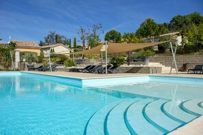 A clear swimming pool with sun loungers and a shaded area, surrounded by a stone wall and greenery.