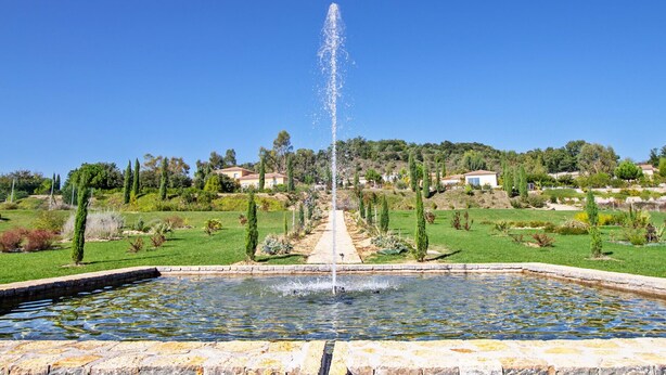A fountain in a garden with a clear sky, surrounded by greenery and houses in the background.