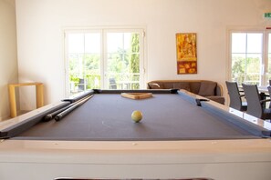 A pool table with a cue ball, a sofa, and a chair in a room with large windows.