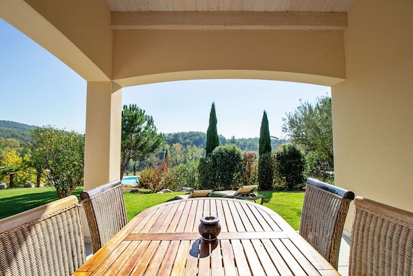A covered outdoor seating area with a wooden table and wicker chairs, overlooking a landscaped garden and distant hills.
