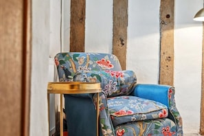Une chaise recouverte d’un tissu à motifs floraux et une petite table d’appoint dorée, placées contre un mur blanc orné de poutres en bois.