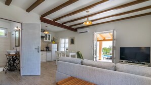 A modern living room with a sofa, wooden table, and a TV. Adjacent is a kitchen with white cabinets and a dining area.