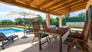 An outdoor patio with a wooden table and chairs, a pool, and a covered area with a view of the surrounding landscape.