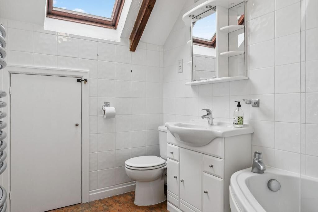 Une salle de bain avec des toilettes, un lavabo et une baignoire, des carreaux blancs et une lucarne.